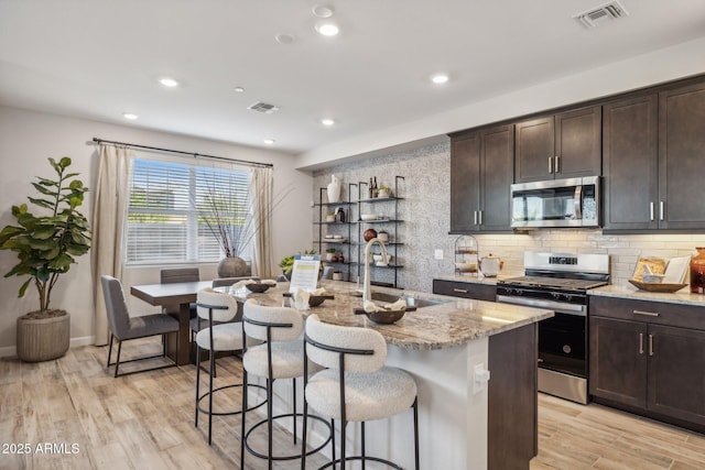 kitchen with sink, a kitchen island with sink, dark brown cabinetry, stainless steel appliances, and light stone countertops