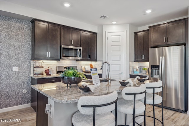 kitchen with stainless steel appliances, dark brown cabinetry, light stone counters, and light hardwood / wood-style flooring