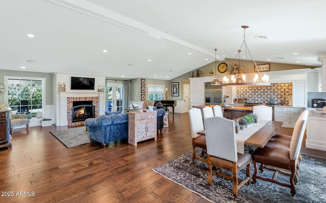 dining area with a brick fireplace, dark wood-type flooring, a chandelier, and vaulted ceiling with beams