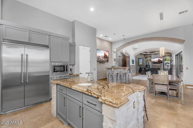 kitchen featuring arched walkways, built in appliances, a sink, gray cabinetry, and light stone countertops
