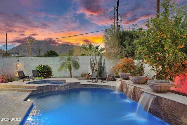 pool at dusk featuring a pool with connected hot tub, a patio, fence, and a mountain view