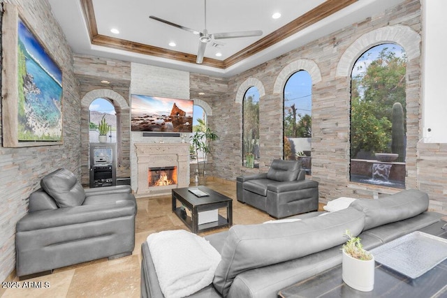 living area featuring a lit fireplace, a ceiling fan, stone tile flooring, and crown molding