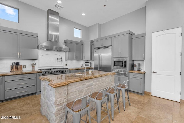 kitchen with built in appliances, wall chimney range hood, a high ceiling, gray cabinetry, and light stone countertops
