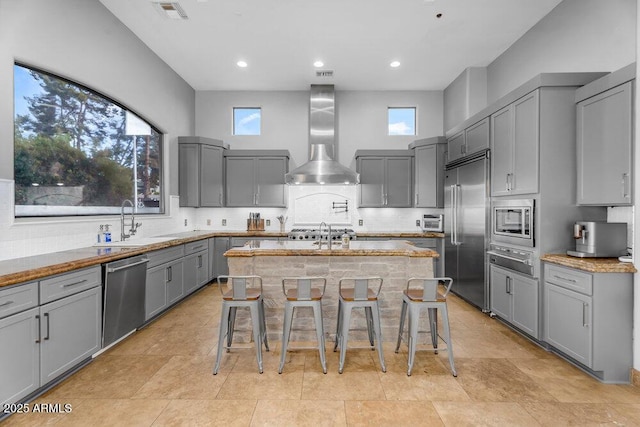 kitchen with visible vents, wall chimney exhaust hood, built in appliances, an island with sink, and gray cabinets