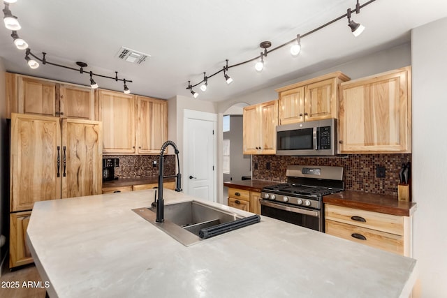 kitchen featuring stainless steel appliances, a sink, light brown cabinetry, decorative backsplash, and arched walkways