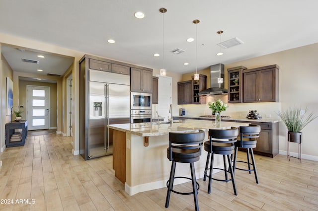 kitchen featuring light stone countertops, wall chimney exhaust hood, built in appliances, an island with sink, and pendant lighting