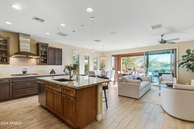 kitchen featuring light stone counters, ceiling fan, sink, wall chimney range hood, and an island with sink