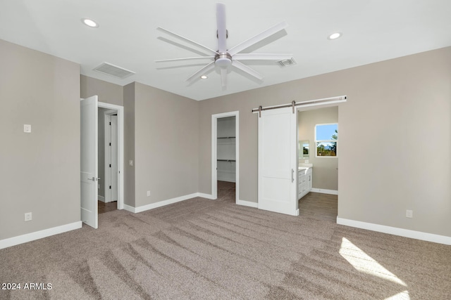 unfurnished bedroom featuring ensuite bath, a spacious closet, a barn door, light colored carpet, and ceiling fan