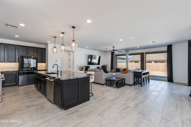 kitchen with sink, an island with sink, stainless steel appliances, decorative light fixtures, and light stone countertops