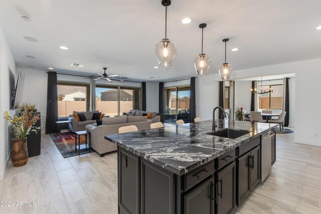 kitchen with sink, stainless steel dishwasher, a center island with sink, dark stone counters, and ceiling fan with notable chandelier