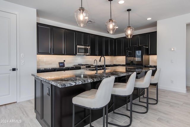 kitchen featuring decorative light fixtures, a center island with sink, backsplash, black refrigerator with ice dispenser, and dark stone counters