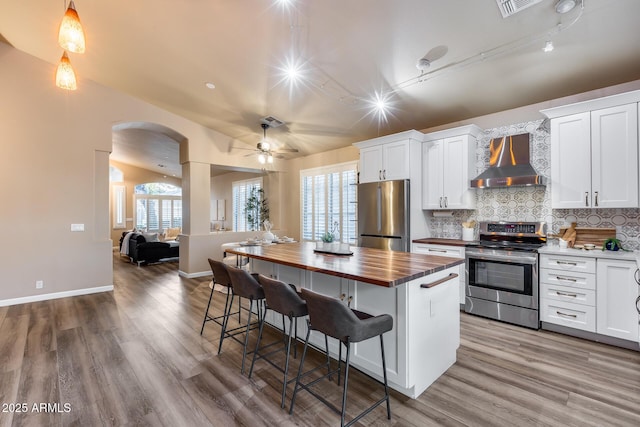 kitchen featuring white cabinets, appliances with stainless steel finishes, a center island, wall chimney range hood, and wooden counters