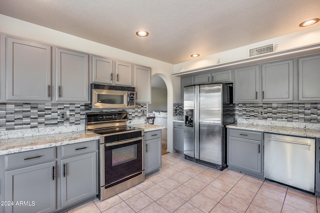 kitchen featuring decorative backsplash, light tile patterned flooring, gray cabinets, appliances with stainless steel finishes, and light stone counters