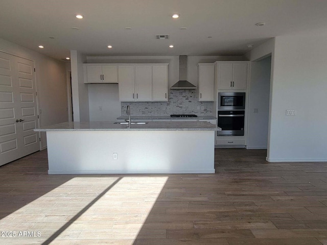 kitchen featuring wall chimney exhaust hood, tasteful backsplash, an island with sink, oven, and white cabinets