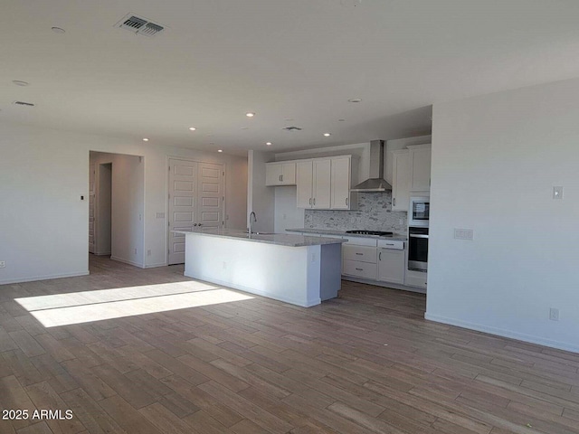 kitchen featuring sink, wall chimney exhaust hood, stainless steel appliances, an island with sink, and white cabinets