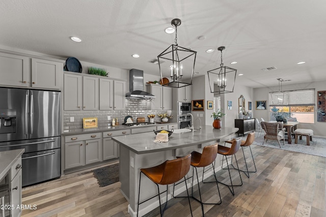 kitchen with appliances with stainless steel finishes, gray cabinetry, wall chimney exhaust hood, hanging light fixtures, and a kitchen island with sink