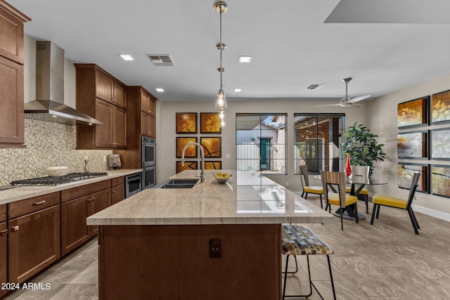 kitchen featuring wall chimney exhaust hood, stainless steel appliances, sink, decorative light fixtures, and an island with sink