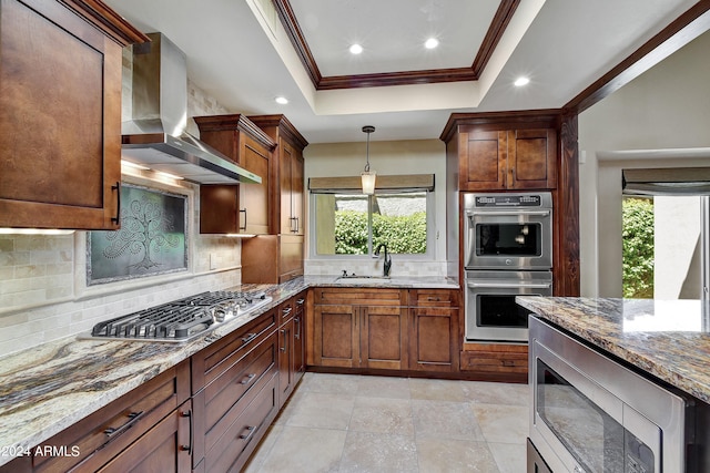 kitchen featuring wall chimney exhaust hood, decorative light fixtures, a tray ceiling, backsplash, and stainless steel appliances