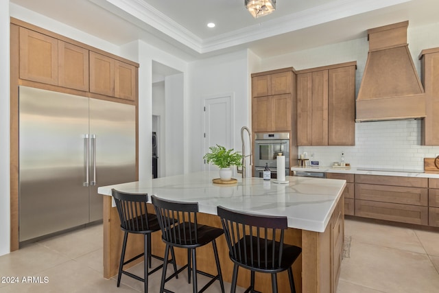 kitchen with custom exhaust hood, appliances with stainless steel finishes, light tile patterned floors, and a kitchen island with sink