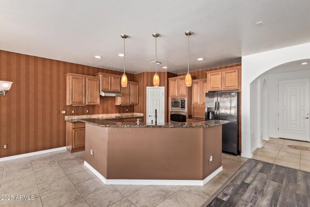 kitchen featuring dark stone countertops, a kitchen island, under cabinet range hood, appliances with stainless steel finishes, and arched walkways