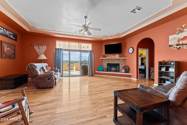 living room featuring ceiling fan, a stone fireplace, a tray ceiling, light hardwood / wood-style flooring, and a textured ceiling