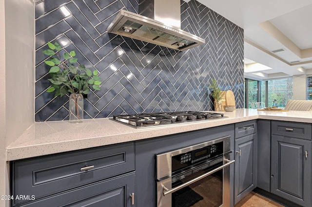 kitchen featuring wall chimney range hood, light stone countertops, decorative backsplash, gray cabinets, and appliances with stainless steel finishes