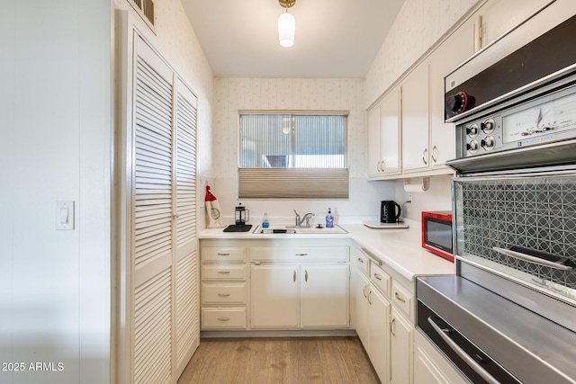 kitchen featuring wallpapered walls, light countertops, light wood-style floors, white cabinetry, and a sink