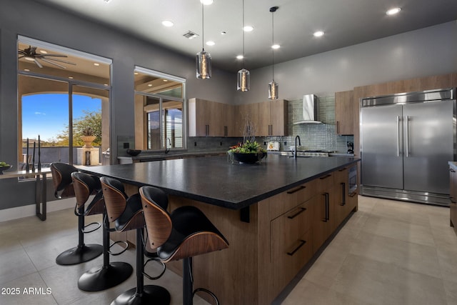 kitchen featuring a kitchen breakfast bar, stainless steel built in fridge, wall chimney exhaust hood, decorative light fixtures, and a center island