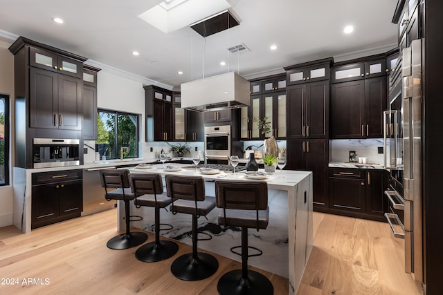 kitchen with a kitchen island with sink, light wood-type flooring, ornamental molding, hanging light fixtures, and appliances with stainless steel finishes