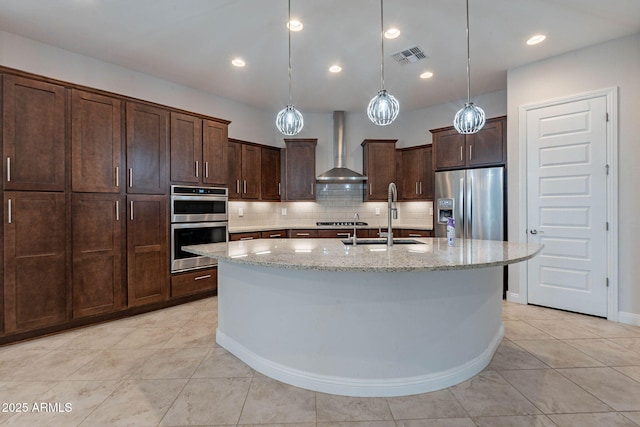 kitchen featuring stainless steel appliances, a sink, light stone countertops, wall chimney exhaust hood, and an island with sink