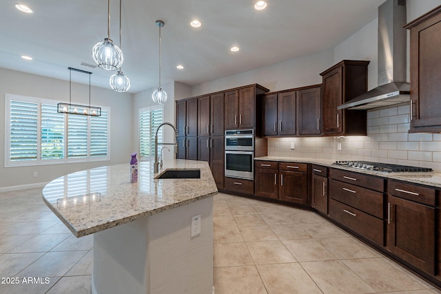 kitchen featuring light stone counters, a sink, appliances with stainless steel finishes, wall chimney exhaust hood, and a center island with sink