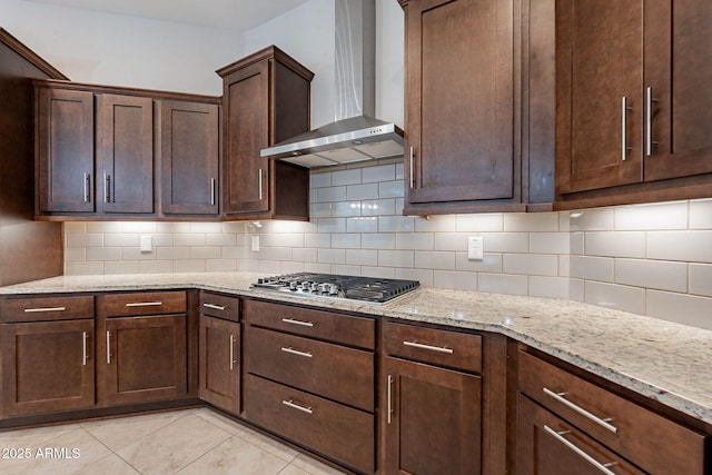 kitchen featuring dark brown cabinetry, wall chimney exhaust hood, light stone counters, stainless steel gas cooktop, and backsplash