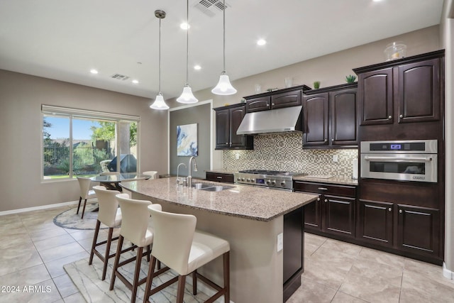 kitchen with light stone counters, a center island with sink, stainless steel appliances, hanging light fixtures, and sink