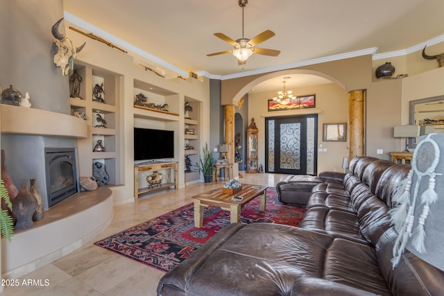 living room featuring built in shelves, a fireplace, arched walkways, ornamental molding, and ceiling fan with notable chandelier