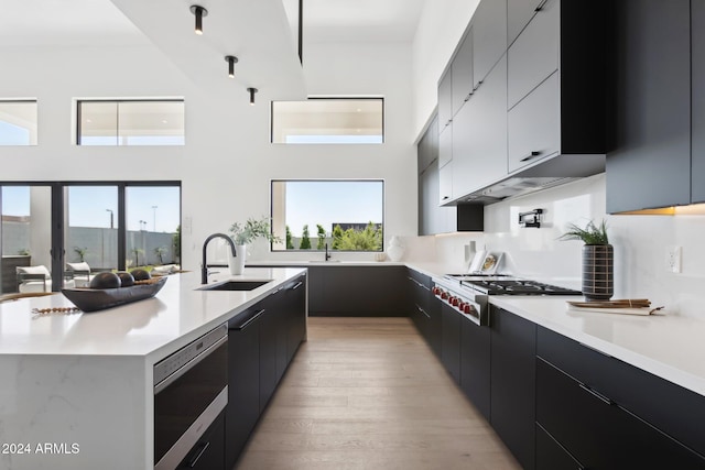 kitchen with light wood-type flooring, a kitchen island with sink, stainless steel gas cooktop, sink, and a high ceiling