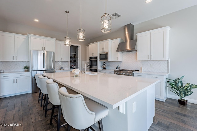 kitchen with wall chimney exhaust hood, white cabinetry, decorative light fixtures, a center island with sink, and stainless steel fridge