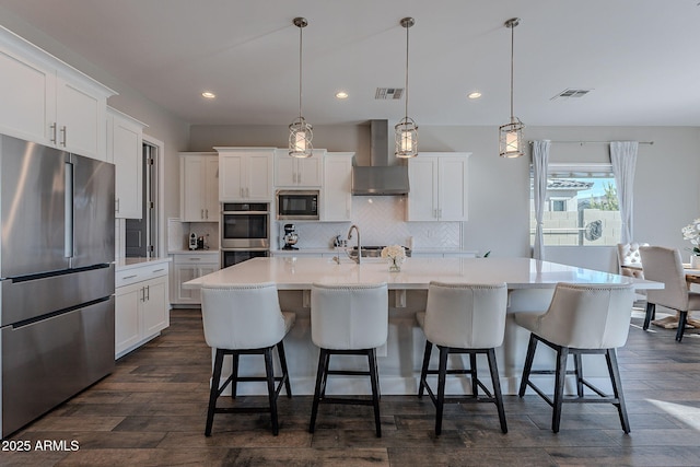 kitchen with white cabinetry, an island with sink, pendant lighting, stainless steel appliances, and wall chimney range hood