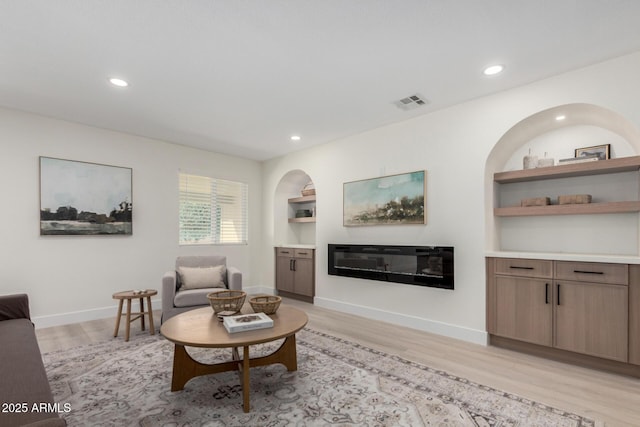 living area featuring baseboards, visible vents, a glass covered fireplace, light wood-style floors, and built in shelves