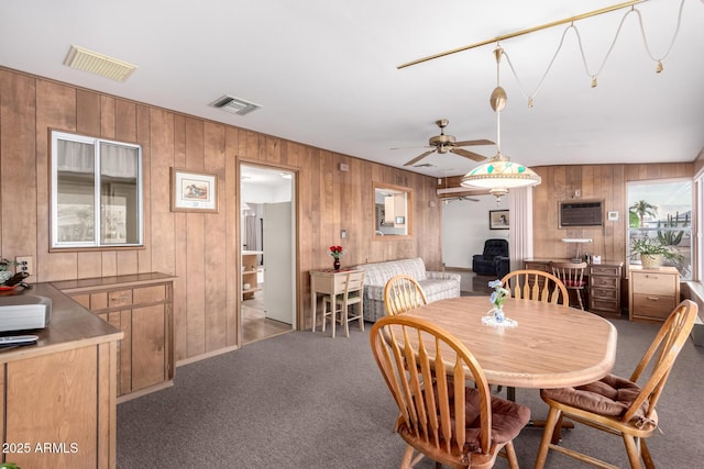 carpeted dining room with a wall mounted AC, ceiling fan, visible vents, and wooden walls