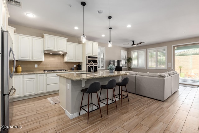 kitchen featuring ceiling fan, decorative light fixtures, an island with sink, appliances with stainless steel finishes, and white cabinetry