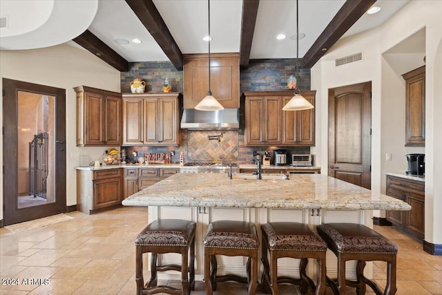 kitchen featuring beam ceiling, hanging light fixtures, a kitchen breakfast bar, a kitchen island with sink, and light stone counters