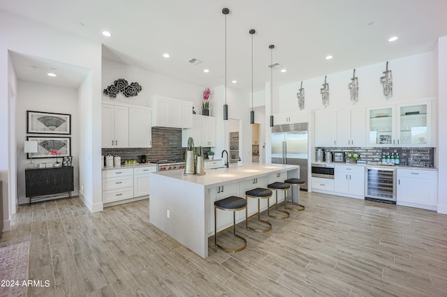 kitchen featuring backsplash, stainless steel appliances, white cabinetry, wine cooler, and an island with sink