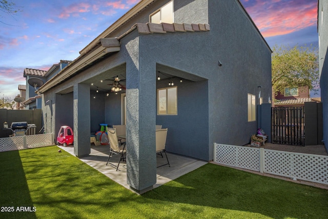 back of property at dusk with a tile roof, a patio, stucco siding, a lawn, and fence