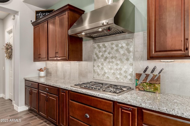 kitchen with wood finished floors, wall chimney range hood, baseboards, decorative backsplash, and stainless steel gas cooktop