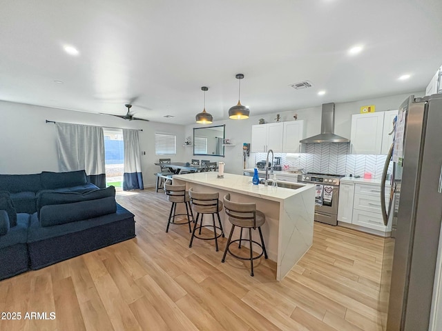 kitchen featuring wall chimney exhaust hood, stainless steel appliances, decorative light fixtures, a center island with sink, and white cabinetry