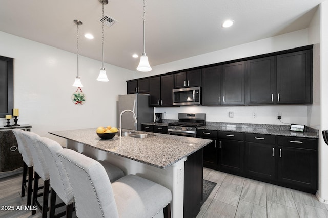 kitchen featuring sink, appliances with stainless steel finishes, a kitchen island with sink, light stone countertops, and decorative light fixtures