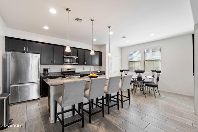 kitchen with appliances with stainless steel finishes, decorative light fixtures, a breakfast bar area, a kitchen island with sink, and light stone countertops