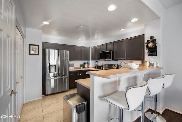 kitchen featuring a kitchen bar, stainless steel appliances, kitchen peninsula, light tile patterned flooring, and dark brown cabinets