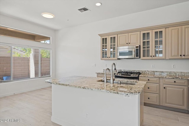 kitchen featuring a center island with sink, glass insert cabinets, stainless steel microwave, light stone counters, and a sink