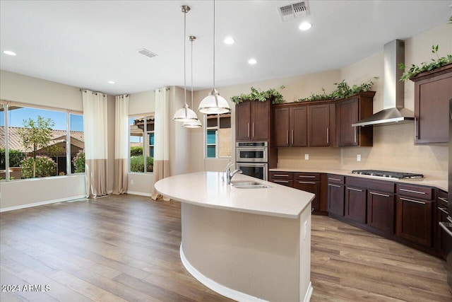 kitchen featuring an island with sink, wall chimney range hood, appliances with stainless steel finishes, hardwood / wood-style floors, and decorative light fixtures